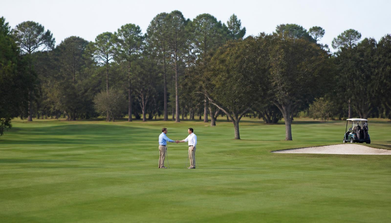 Two golfers in business-casual attire shaking hands on a fairway